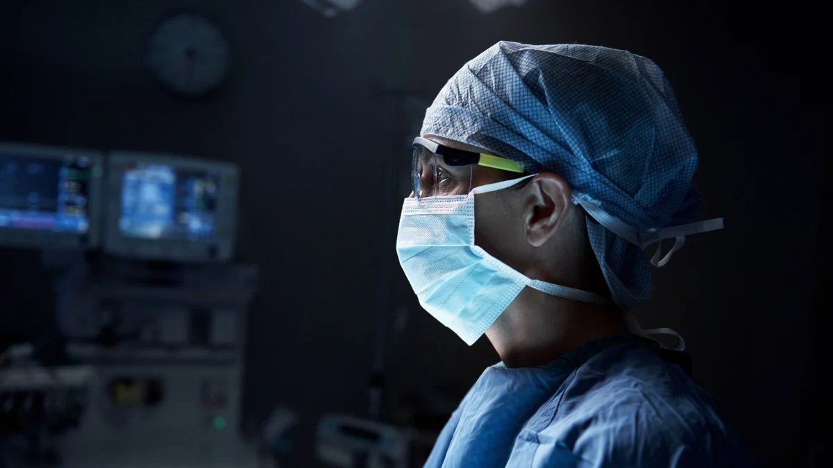 A clinician in a surgical mask and cap in a dimly lit hospital room.